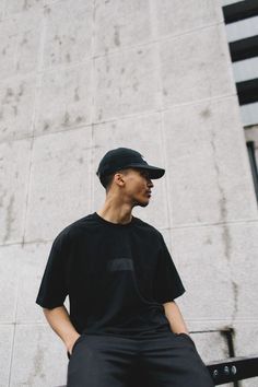 a man sitting on a bench in front of a building wearing a black shirt and cap