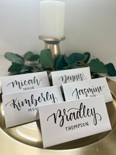 four place cards with names on them sitting on a gold plate next to a candle