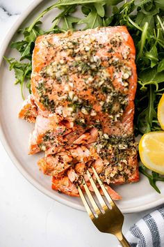 a white plate topped with salmon next to a pile of greens and lemon wedges