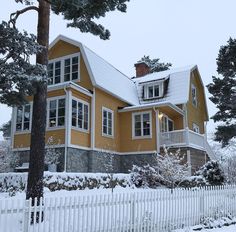 a yellow house with snow on the ground and trees around it, in front of a white picket fence