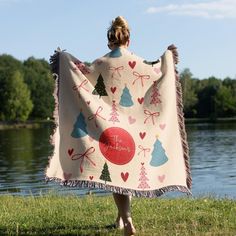 a woman is holding up a blanket with christmas trees and bows on it in front of a lake