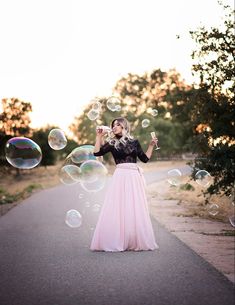 a woman in a long pink skirt blowing bubbles on the road with trees and bushes behind her