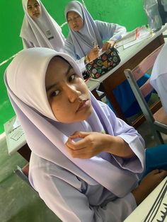 two women in hijabs are sitting at a desk and one is eating something