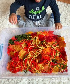 a young boy sitting on the floor in front of a plastic container filled with fall leaves