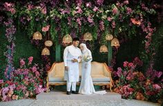 two people standing next to each other in front of a flower covered wall with flowers on it