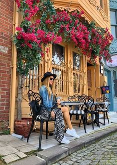 a woman sitting on a bench in front of a building with flowers hanging from it