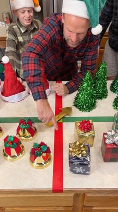 a man cutting a red ribbon on top of a table with christmas decorations and presents