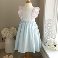 This Little Girl's Pinafore Dress Is Perfect For Parties, Especially Tea Parties! The Cotton Blend Dress Makes For Easy Care, Less Wrinkles. The Base Of The Dress Is Done In A Beautiful Aqua Blue. The Bodice Is White And Has A Lovely Hand-Embroidered Floral Wreath Done Is Pastel, Blue, Green, Yellow And Pink. The Flowers Are Done Bullion Style. Pink Ruffles Attach To The Bodice And Over The Shoulders And Down The Top Of The Back. The Ruffle Is Trimmed In A Hand-Embroidered Matching Aqua Blue Sca Easter Dresses With Ruffles And Short Sleeves, Light Blue Sleeveless Dress For Baptism, Fitted Light Blue Princess Dress For Spring, Cute Sleeveless Light Blue Princess Dress, Blue Ruffled Princess Dress For Summer, Light Blue Ruffled Princess Dress For Baptism, Blue Summer Princess Dress With Ruffles, Fitted Princess Style Easter Dresses, Fitted Princess Dresses For Easter