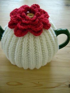 a knitted tea cosy with a red flower on the top sits on a wooden table