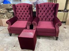 two red velvet chairs with matching footstools in a warehouse setting, one has a stool and the other has a small end table