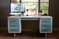 a desk with a computer on it in front of a window and a potted plant