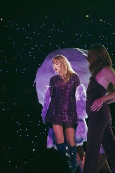 two women standing on stage in front of a large object with purple feathers and sequins