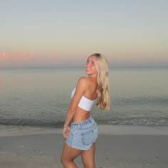 a beautiful young blond woman standing on top of a sandy beach next to the ocean