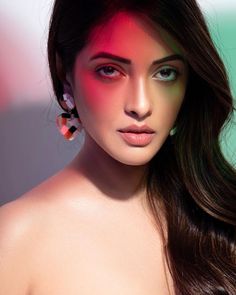 a woman with long dark hair and bright makeup looks at the camera while wearing earrings