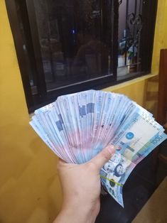 a person holding up several bills in front of a window with a yellow wall behind them