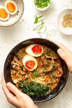 two hands holding a bowl of food with eggs and spinach on the table next to other dishes