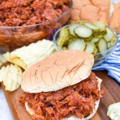 a pulled pork sandwich with pickles and potato chips on a wooden cutting board next to a bowl of pickles