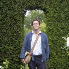 a man standing in front of a green hedge with his hand on his hip and looking at the camera