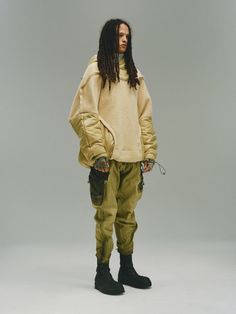 a man with dreadlocks standing in front of a gray background wearing an off white sweater and khaki cargo pants