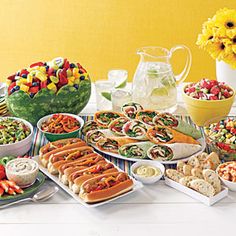 a table full of food including salads, sandwiches and desserts with flowers in the background