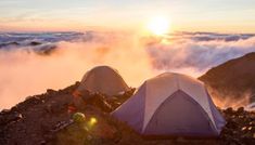 two tents set up on top of a mountain with the sun setting in the background