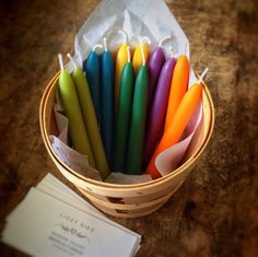 a basket filled with lots of colorful candles next to a white envelope on top of a wooden table