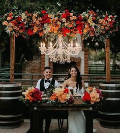 a man and woman sitting at a table in front of wine barrels with chandelier