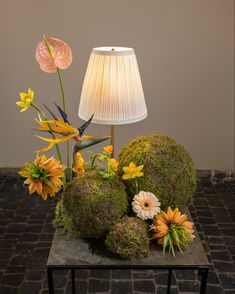 an arrangement of flowers and moss sits on a table with a lamp in the background