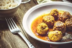 a white bowl filled with meatballs and gravy next to rice on a wooden table