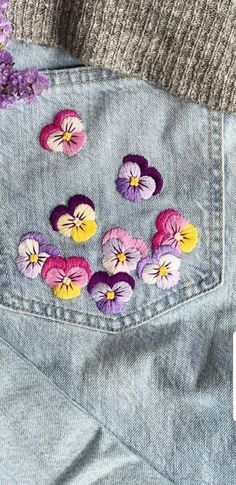 a pair of jeans with flowers embroidered on the back and side pocket, sitting next to a purple flower