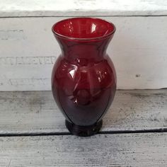 a red glass vase sitting on top of a wooden table next to a white wall