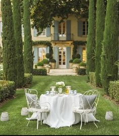 an outdoor table set for two in front of a large house with trees and bushes