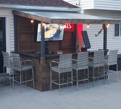 an outdoor bar with six chairs and a television on the back wall, in front of a white house