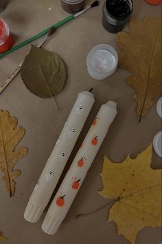 two white candles sitting on top of a table next to autumn leaves and paintbrushes