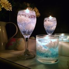 two wine glasses with water and ice in them sitting on a table next to some tea lights