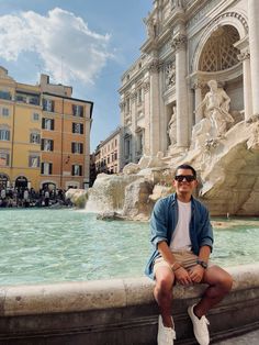 a man sitting on the edge of a fountain in front of some buildings and water