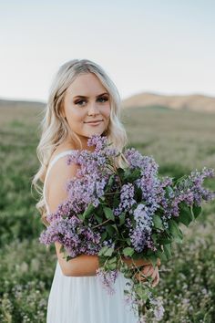 a woman in a white dress holding a bouquet of purple flowers and smiling at the camera