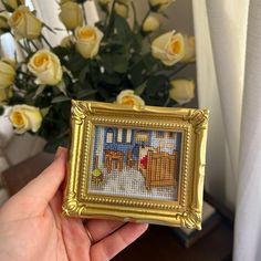 a person holding up a cross - stitch picture in front of a bouquet of roses