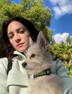 a woman holding a small white dog in her arms and looking at the camera with trees behind her