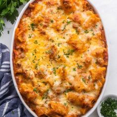 a casserole dish with cheese and parsley on the side next to a small bowl of green herbs