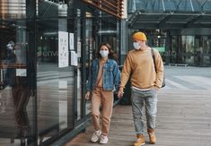 two people wearing face masks walking down the sidewalk in front of a building with glass doors