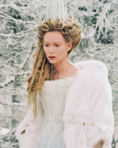 a woman with long blonde hair wearing a white dress and fur coat in front of snow covered trees