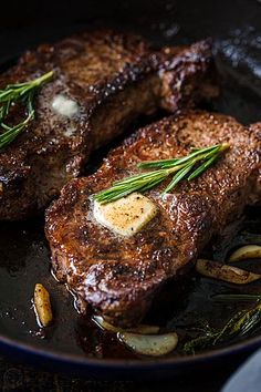 two steaks are being cooked in a skillet with onions and herbs on the side