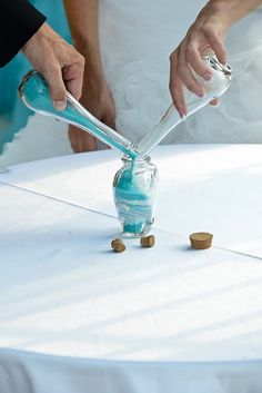 a person pouring something into a glass vase on top of a white cloth covered table