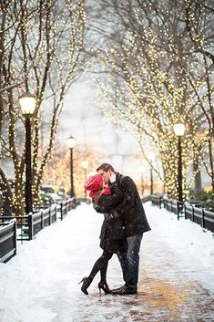 a man and woman kissing in the snow on a street with christmas lights behind them