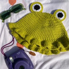 a crocheted frog hat, sunglasses and camera on a bed