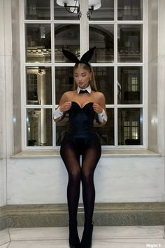 a woman in bunny ears is standing by a window with her legs crossed and looking at the camera