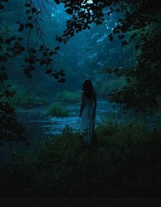 a woman standing in the woods at night with her back turned to the camera, looking out over a body of water