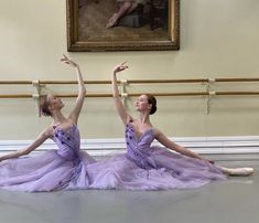 two ballerinas in purple tulle skirts are sitting on the floor with their arms stretched out