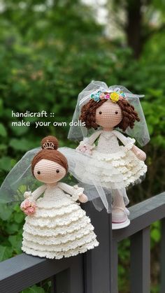 two little dolls dressed in white dresses and veils on a wooden fence with trees behind them